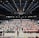 An indoor sports arena filled with rows of tables, each occupied by people engaged in competitive activities. The ceiling is high, with numerous lights illuminating the large space. The audience seating on either side is empty, and there are flags hanging from the upper level.
